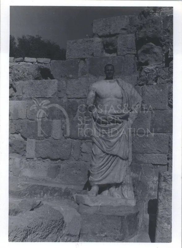 Man standing behind a draped marble statue in front of a stone wall of ancient blocks