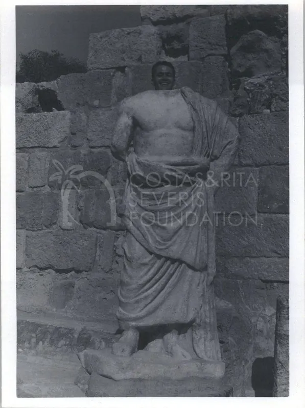 A smiling man's head appears above a draped marble statue in front of a stone block wall