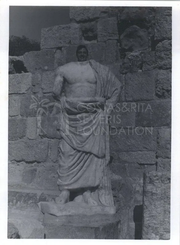 A smiling man's head rises behind a draped marble statue standing against a wall of large stone blocks.