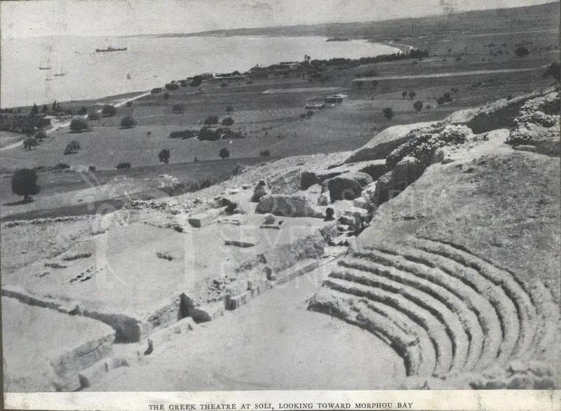 Curved stone seating of a ruined theatre on a hillside, with a bay and boats beyond