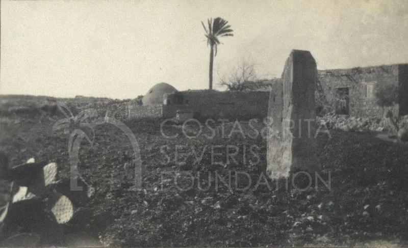 Dusty field with a tall upright stone slab, low walls, a domed structure and a palm tree behind