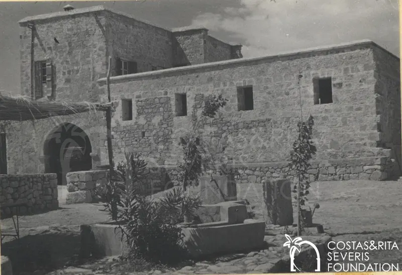 Stone building with an arched doorway and small windows, with a stone trough and plants in the yard