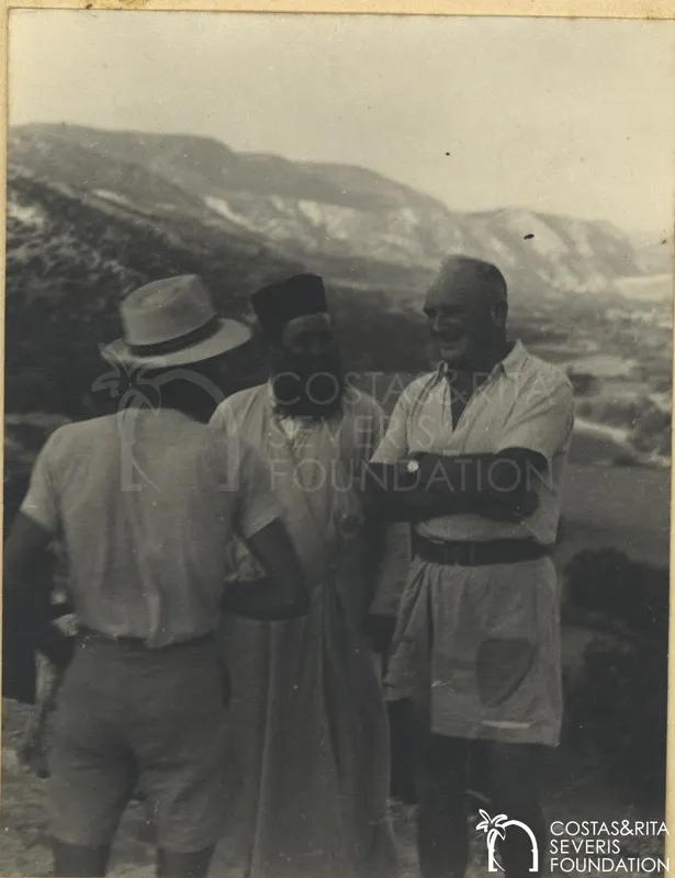 Three men talking on a hillside, one in a dark cap and robe, with mountains behind them