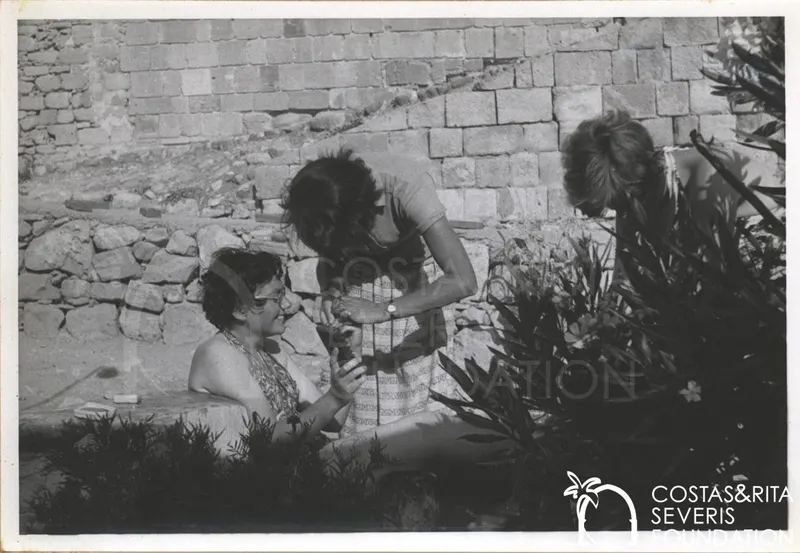 Three women outdoors beside a stone wall and oleander bushes, one seated and one bending toward her