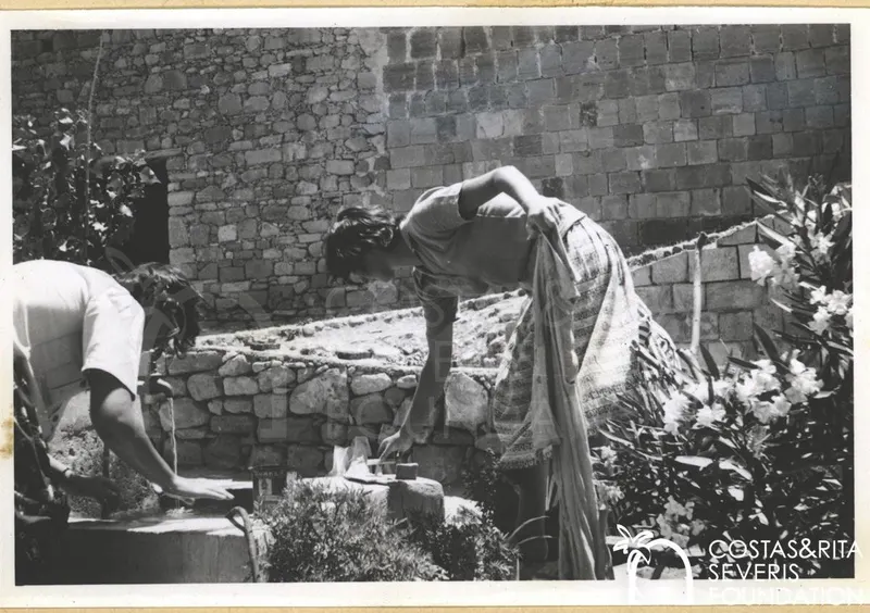 Two people bending over a stone basin with running water beside a rough stone wall and oleanders