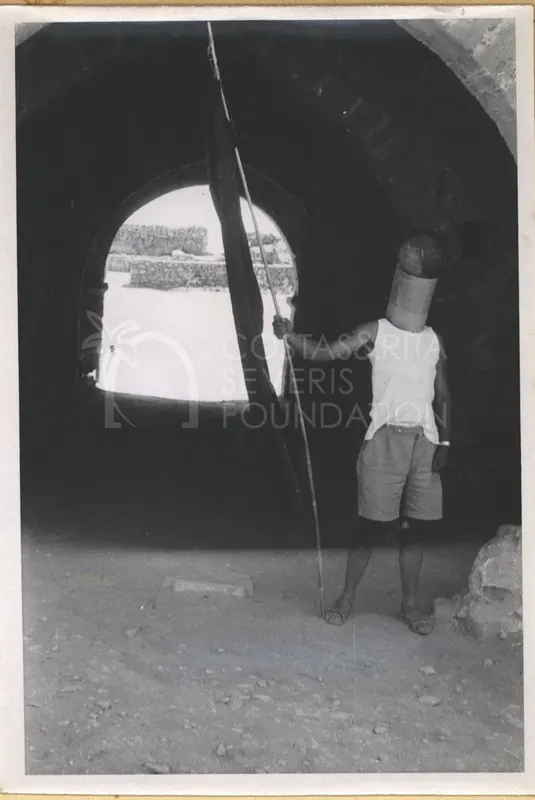 Man in a cylindrical helmet and white vest holding a pole with a dark flag, standing under a stone archway
