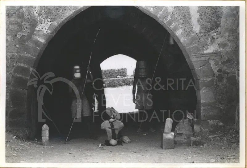 Two men in improvised costumes stand under a stone archway, one holding a long pole