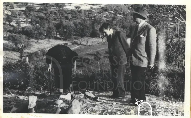 Woman bending over stones on a hillside, with two men in suits and hats standing beside her