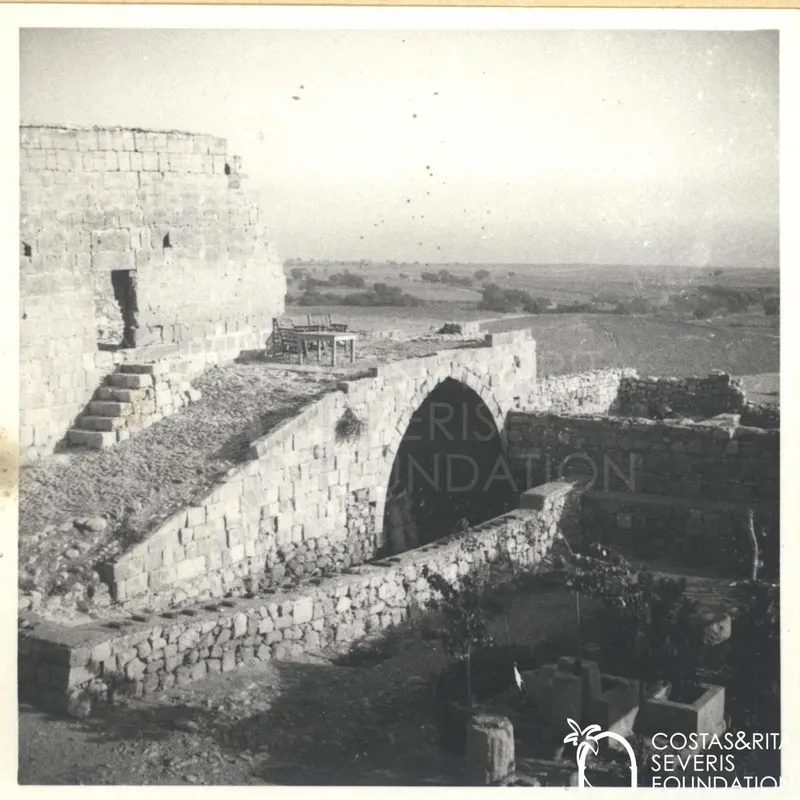 Stone ruins with an arch, steps and a wooden table on a terrace, with open fields beyond