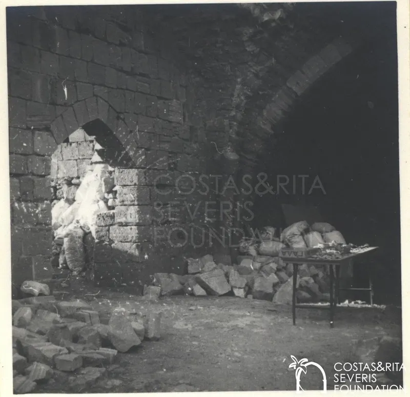 Vaulted stone chamber with sacks and cut stone blocks on the floor and a table of fragments at right
