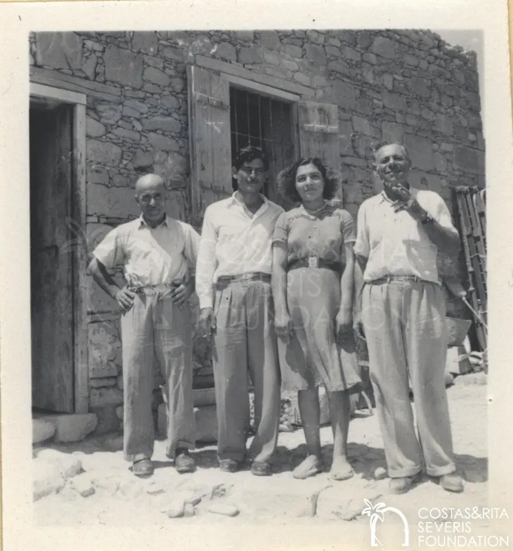 Four people stand side by side in front of a stone building with a wooden door and barred window