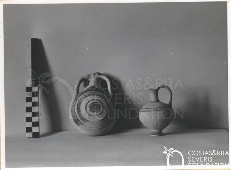 Two clay vessels on a surface beside a checkered scale rod against a grey background