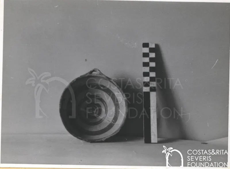 Shallow pottery vessel with a small loop handle beside a checkered scale rod