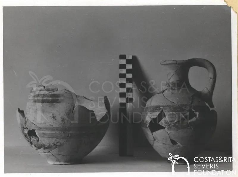 Two broken pottery vessels beside a chequered scale rod against a grey backdrop