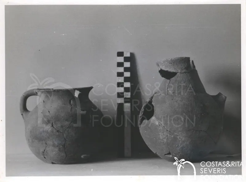 Two broken pottery vessels, a handled jug on the left and a larger jar on the right, with a chequered scale rod between them