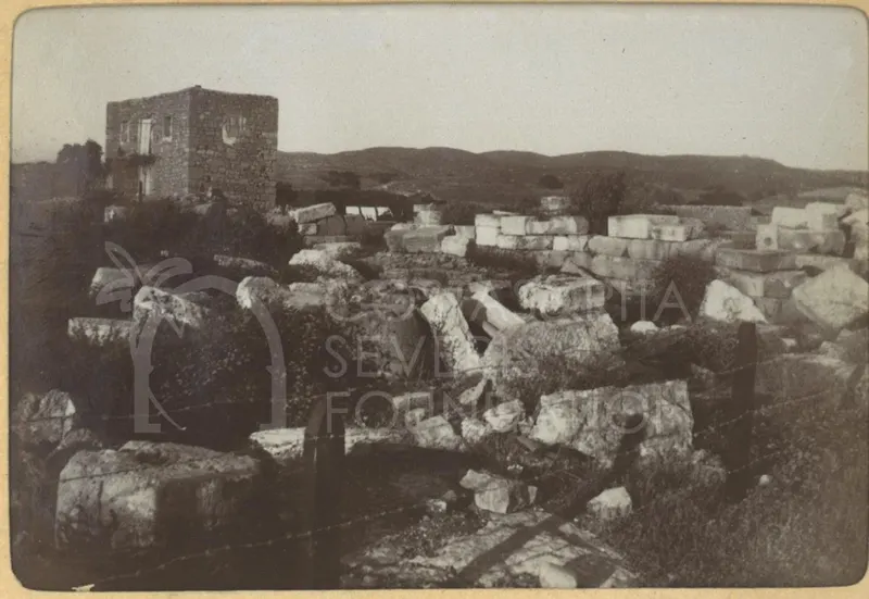 Scattered cut stone blocks among scrub, with a stone building at left and hills behind