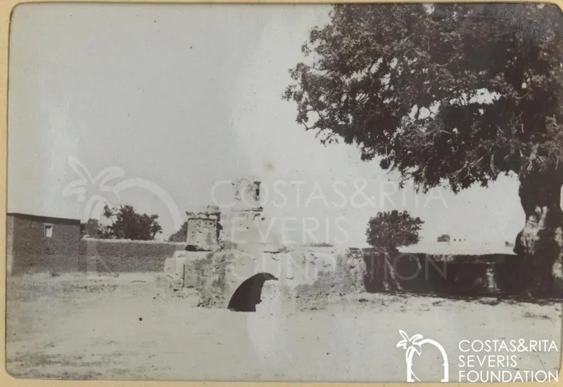 Ruined stone structure with an arched opening on bare ground, with a large tree at right