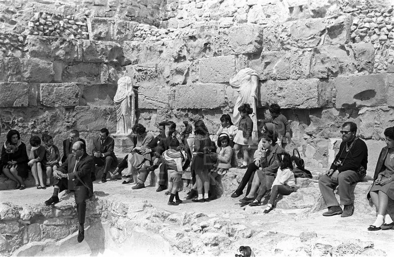 Visitors sit on stone steps before a stone wall at Salamis, with two draped statues beside them.