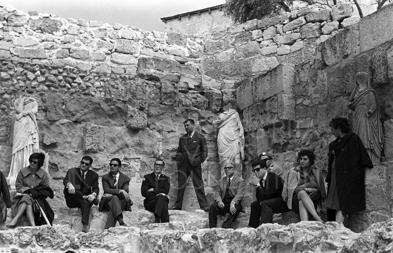 Group of seated and standing people in front of ancient stone walls, with marble statues beside them