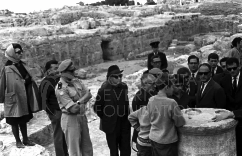 Group of visitors and a uniformed man among ancient stone ruins, with a carved stone block at right