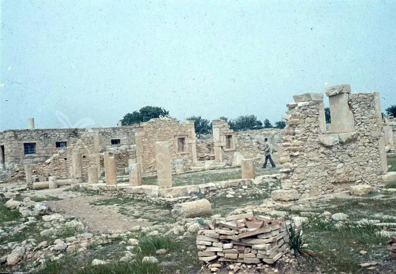 Ruined stone walls and column stumps on a grassy site, with a man walking among them