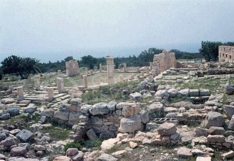 Scattered ancient stone ruins, columns and low walls on a grassy hillside, with trees and hazy sky beyond