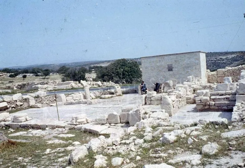 Ruined stone walls and scattered blocks of the Sanctuary of Apollo Hylates, with two seated people and a modern stone building