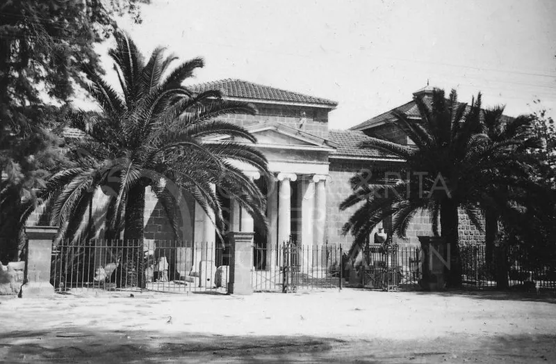 Columned stone building with a pediment and tiled roof behind iron railings, framed by palm trees.