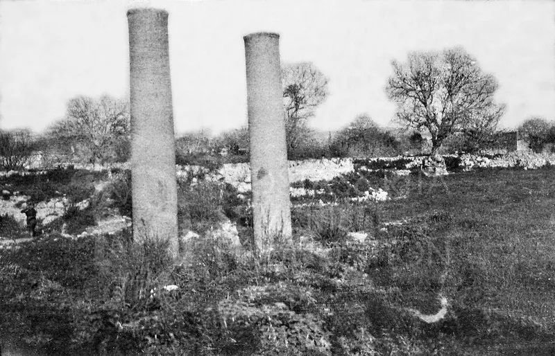 Two tall stone pillars standing in a grassy field among low ruined walls, with bare trees behind