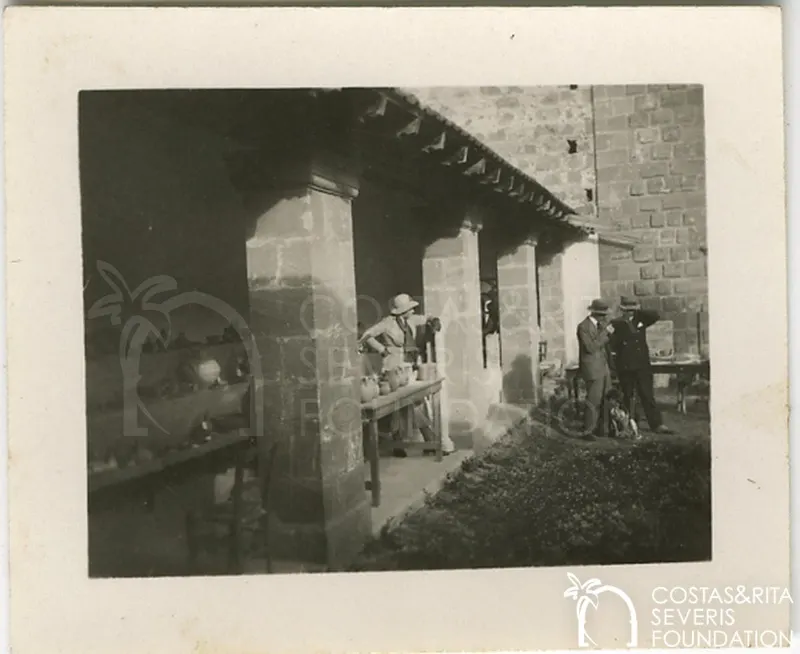 Woman at a table of pottery under a stone arcade, with two men in suits and hats standing nearby