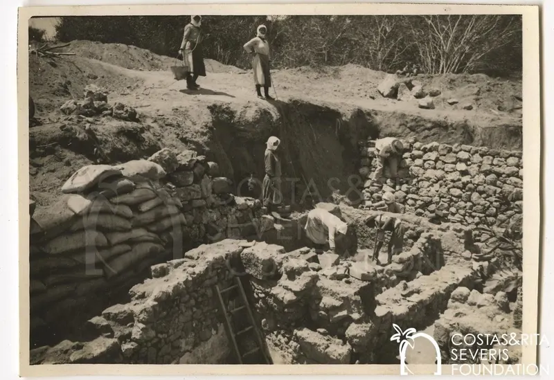 Workers excavating a trench with exposed stone walls, sandbags at left and women standing on the bank above