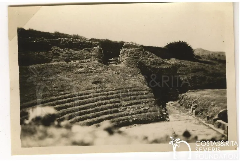 Tiered stone rows of ancient ruins rising on a hillside, with a paved path to the right