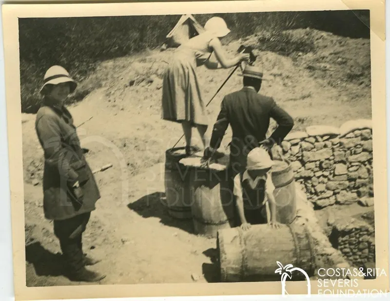 Four people by barrels and a stone wall on a dirt slope, one woman raising an arm as if taking a photograph