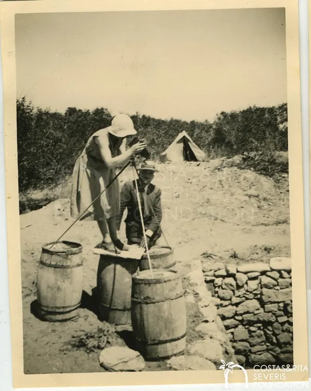 Woman in a pith helmet photographing from barrels on a dry excavation site, with a seated man beside her