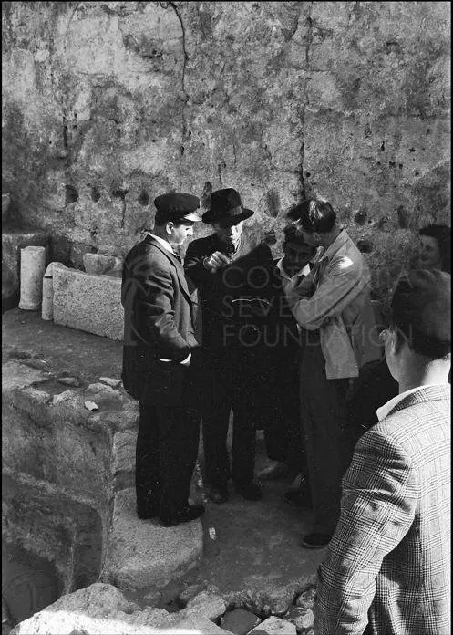 Men gathered in front of a rough stone wall, one in a dark hat speaking to the others