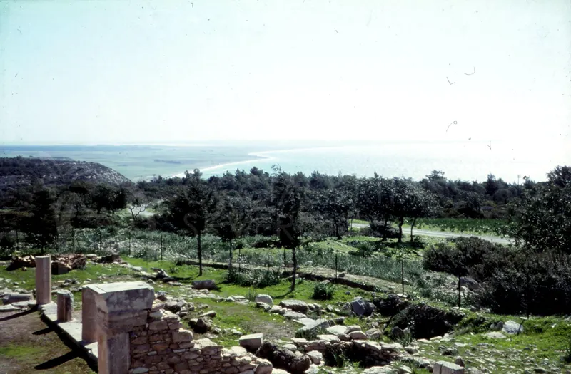 Ruined stone walls and columns in the foreground, with trees and a bay and sea beyond