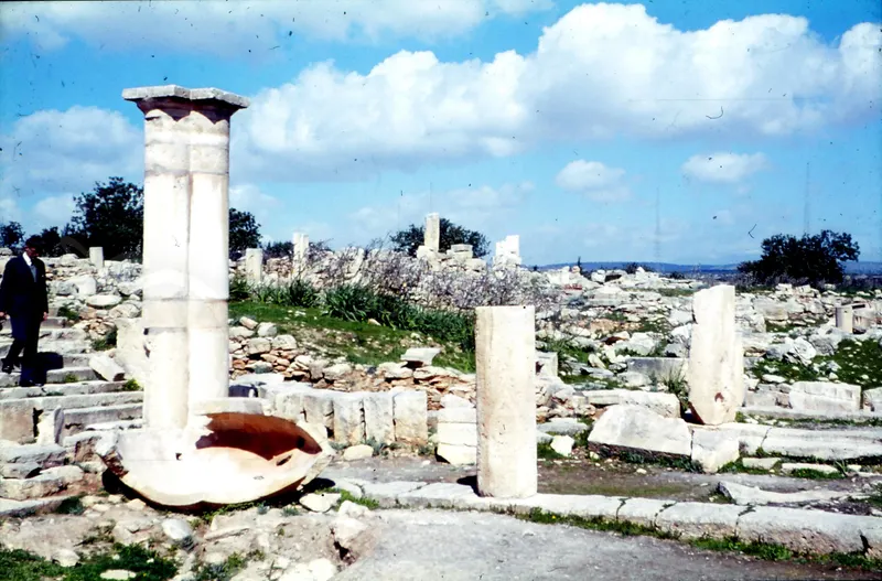 Ruined marble columns and a broken stone basin among scattered masonry, with a man on steps at left