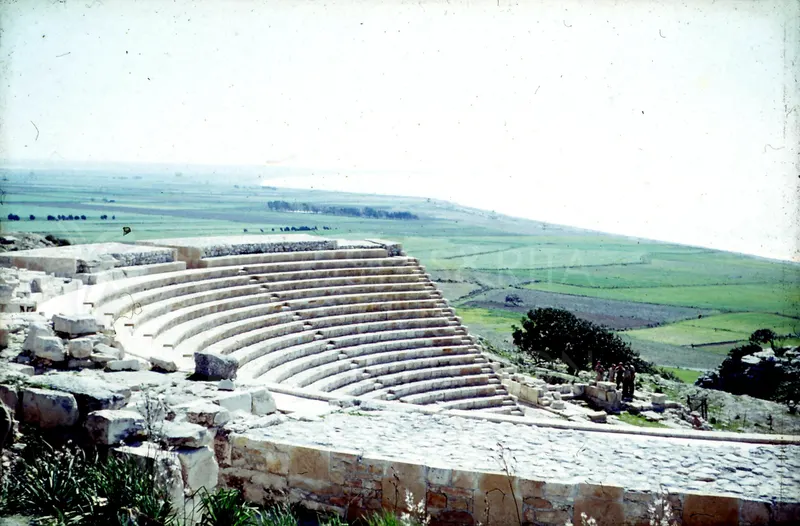 Curved stone tiers of a theatre on a hillside, with green fields and the coast beyond