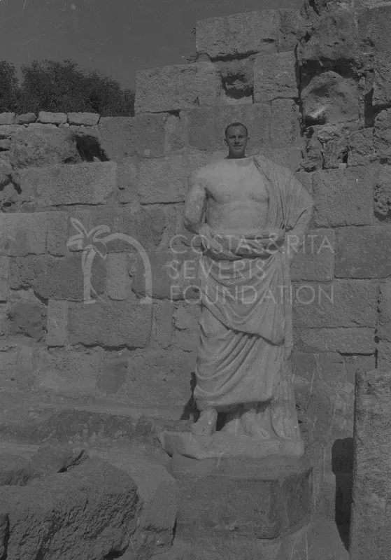 A man's head and shoulders rise behind a headless draped marble statue against a stone block wall