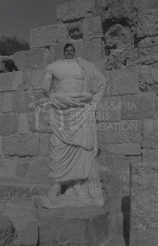 Man smiling behind a draped marble statue against a stone block wall at Salamis