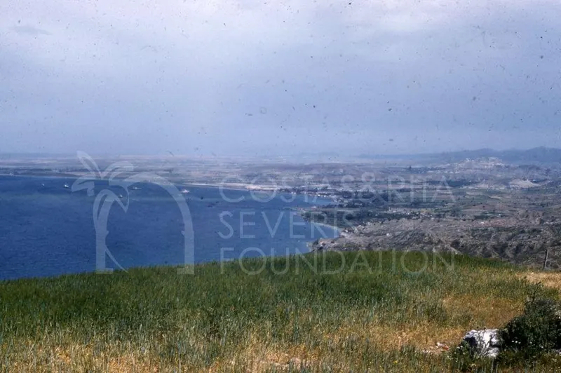 Blue bay curving along a coast, with grassy ground in the foreground and hills and a settlement beyond