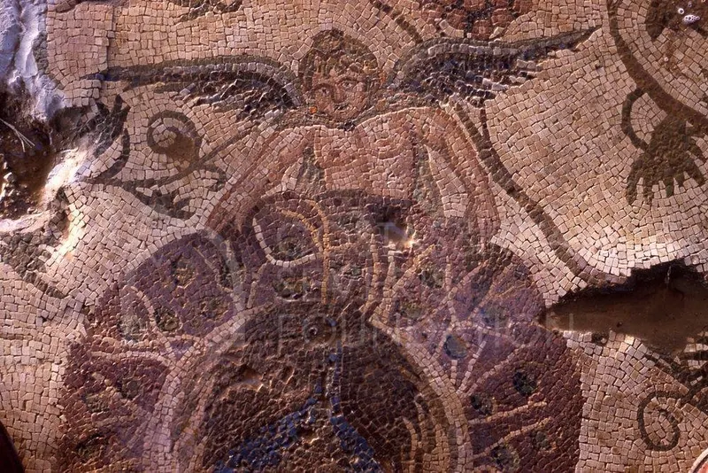 Close view of a mosaic floor showing a winged figure with a face, set among small coloured tesserae