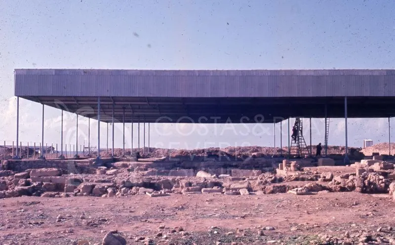 Steel canopy roof over stone wall remains at an excavated site, with figures beside a ladder at right