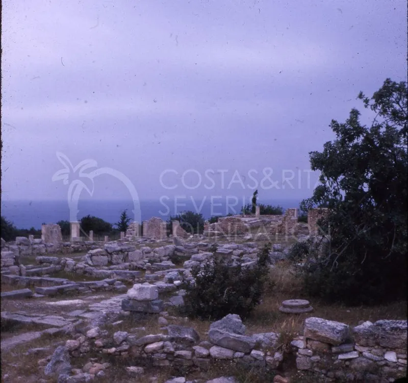 Ruined stone walls and scattered blocks of an ancient site, with the sea on the horizon and a figure standing among the ruins