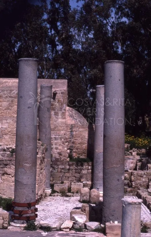 Grey columns stand above a geometric mosaic floor, with a brick wall and dark trees behind them