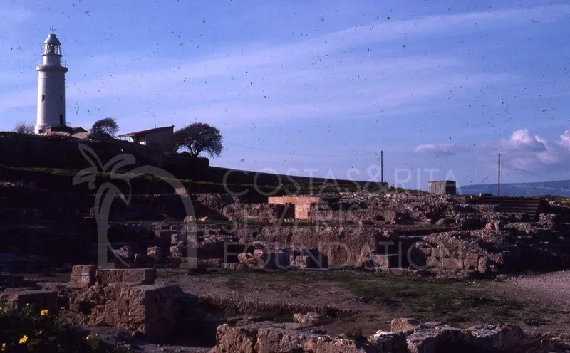 White lighthouse on a hill at left, above stone wall remains and rubble in the foreground
