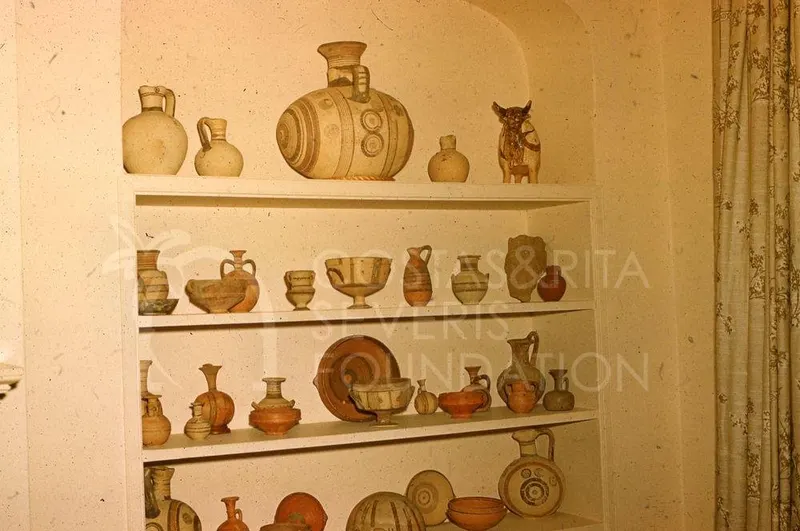 Shelves in a wall niche holding many pottery jugs, bowls and jars, with a small animal figurine at top right