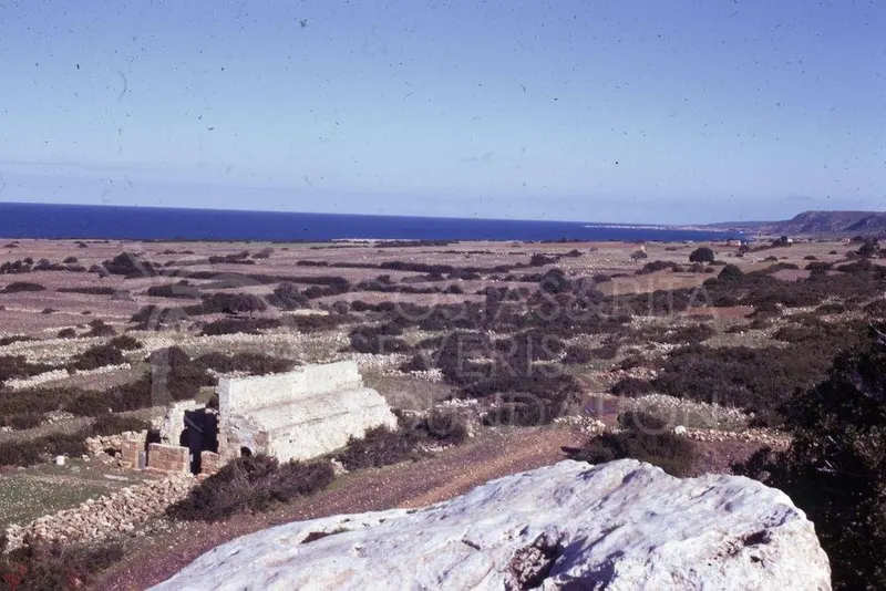 Ruined pale stone structure with an arched opening in scrubland, with the sea on the horizon