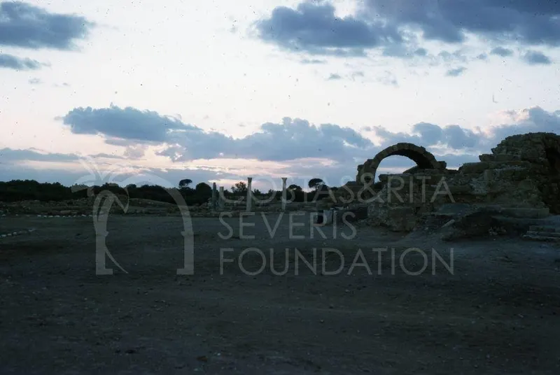 Ruins at dusk with a rounded arch on the right and standing columns in the middle distance under a cloudy sky