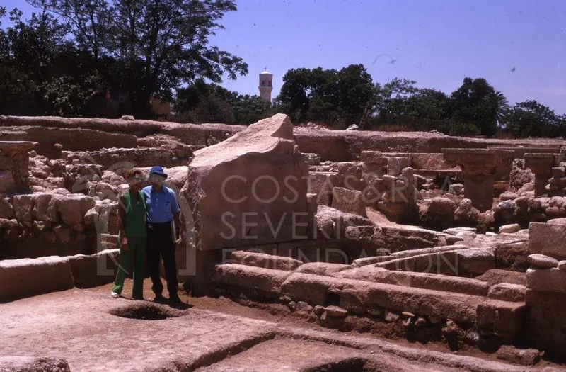 Two people stand among excavated stone ruins with column bases and a large fallen stone block at Kition.
